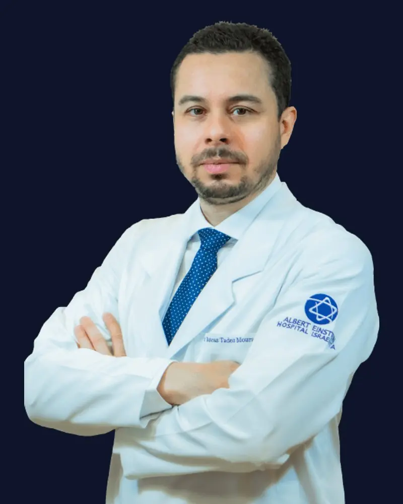 Dr. Lucas Tadeu Moura de jaleco branco e gravata azul, braços cruzados, sobre fundo azul-marinho